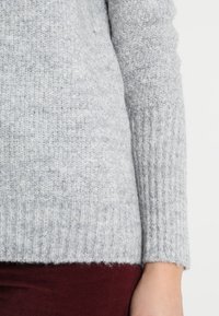 Pull en maille gris clair avec une surface texturée, des poignets et un ourlet côtelés. Matériau doux, ressemblant à de la laine, montré sur la manche contre un fond sombre.