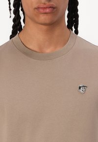 Beige t-shirt lavet af bomuld med rund hals, prydet med et broderet logo af en smilende karakter på venstre brystområde.