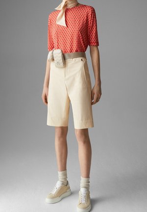 Camicia a maniche corte con fantasia rossa, pantaloni beige e sneakers beige con dettagli bianchi. Una borsa di colore chiaro è attaccata in vita.
