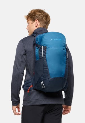 Jeune homme portant un sac à dos Vaude bleu et marine avec des bretelles rembourrées et une ceinture de taille, vêtu d'une veste sombre et d'un pantalon noir, de dos.