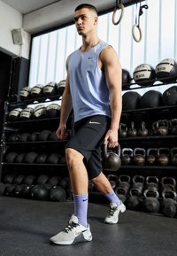 Szara odważnik kettlebell trzymana w prawej ręce; ubrana w jasnoniebieski top bez rękawów, czarne spodenki, fioletowe skarpetki oraz szare buty sportowe z czarnymi akcentami.