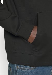 Felpa nera con texture, dotata di tasca a marsupio, polsini a costine e orlo largo. Indossata con jeans blu, che mostra parzialmente la mano.