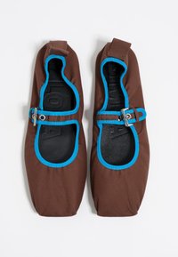 Un par de zapatos de tela marrones tipo slip-on con ribetes de un azul brillante y correas ajustables con hebillas sobre un fondo blanco.