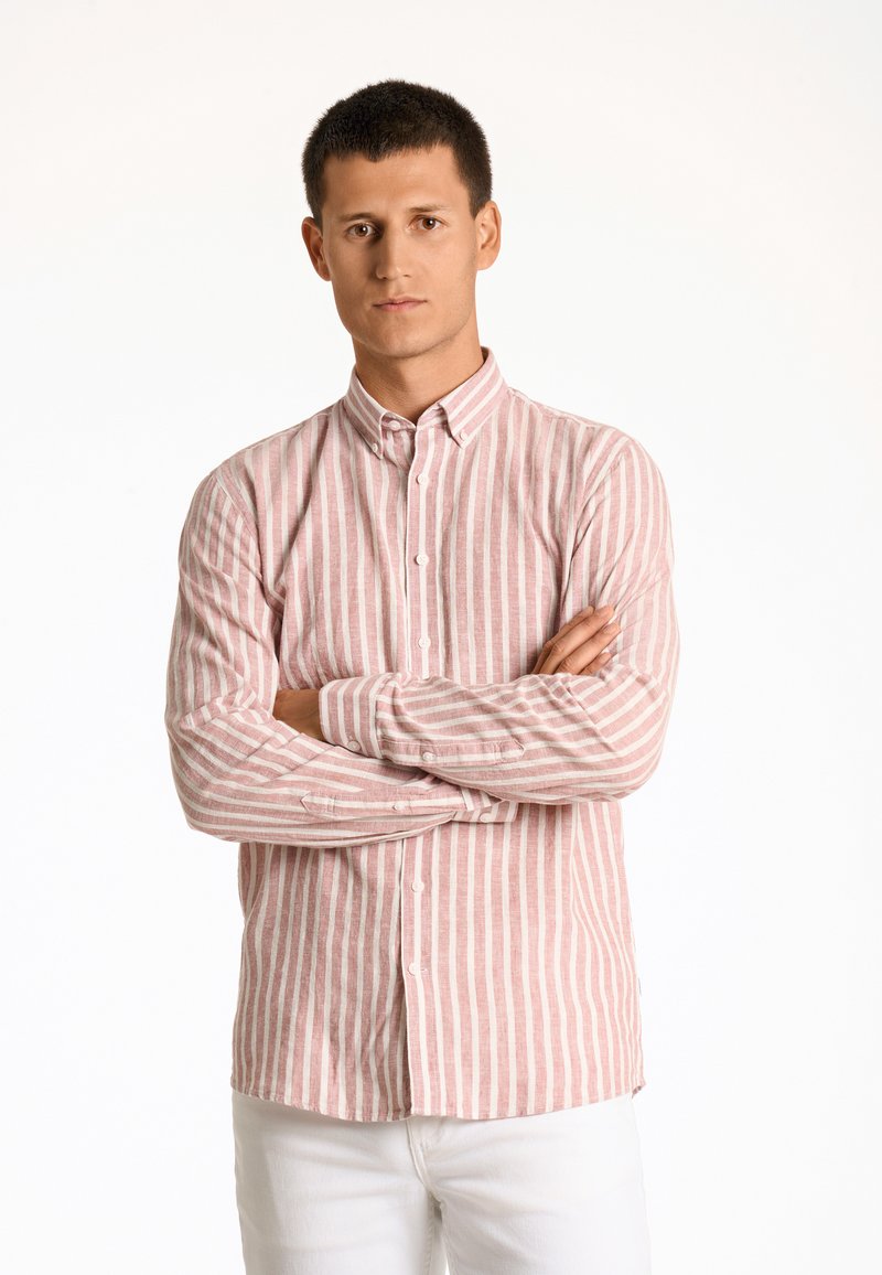 Camicia a righe con bottoni in rosa chiaro e bianco, realizzata in tessuto traspirante, con maniche lunghe e colletto classico. Indossata con pantaloni bianchi.