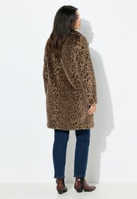Pletteret leopardfrakke i blødt stof, brun med sorte pletter, knælangt design, parret med mørkeblå jeans og brune ankelstøvler.