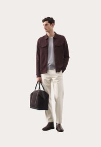 Mørk burgunder jakke med to brystlommer, grå sweater, cremede bukser, brune læderstøvler og en brun duffeltaske med sorte accenter.