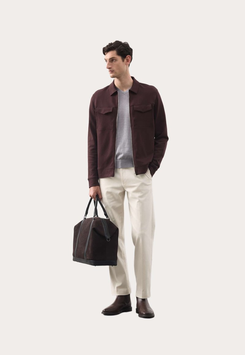 Mørk burgunder jakke med to brystlommer, grå sweater, cremede bukser, brune læderstøvler og en brun duffeltaske med sorte accenter.