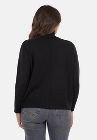 Una donna con lunghi capelli castani che indossa un maglione nero a collo alto e jeans blu, rivolta di spalle contro uno sfondo chiaro e uniforme.