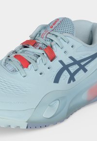 Ανοιχτό μπλε αθλητικό παπούτσι Asics με γκρίζο λογότυπο, αναπνεύσιμη γλώσσα από ίνες και κόκκινες κορδόνια σε λευκό φόντο.