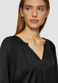 Black blouse with a V-neck, featuring soft fabric, pleated shoulders, and a draped design that falls loosely around the neckline.