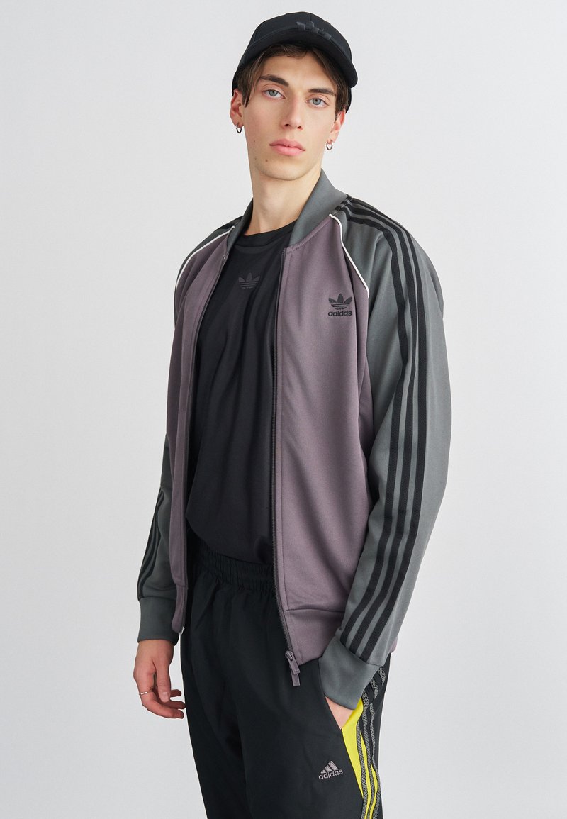 Adidas jakna na zip u sivoj i ljubičastoj boji, sa tri crne pruge duž rukava i crnim logotipom. Nosena preko crne majice.