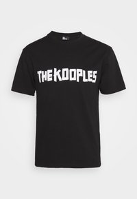 Svart bomullströja med fet vit text som säger "THE KOOPLES" över bröstet. Klassisk rund halsringning och korta ärmar.