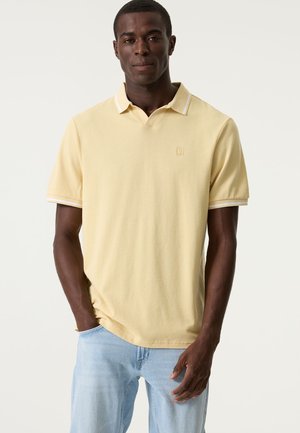 Gele poloshirt met een gestructureerde stof, voorzien van een witte gestreepte kraag en een klein logo op de borst. Gecombineerd met lichtblauwe spijkerbroek.