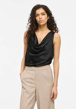 Kobieta o kręconych włosach ubrana w czarny satynowy top bez rękawów z dekoltem typu kopertowego i beżowe spodnie, stojąca z jedną ręką w kieszeni.
