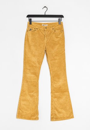 Pantalones de pana de color mostaza con bolsillos delanteros y cierre de botón colgados en una percha negra contra un fondo blanco.