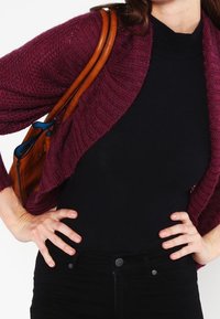 Bordeauxroter Strickcardigan mit offenem Vorderteil und strukturiertem Design, getragen über einem schwarzen, gerippten Top, kombiniert mit einer braunen Lederhandtasche.