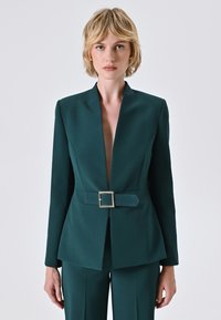 Blazer su misura verde smeraldo con scollatura a V profonda e una cintura larga con fibbia quadrata argentata. Il tessuto appare liscio e lussuoso.