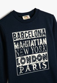 Granatowa bawełniana koszula z długim rękawem z wyraźnym, zniszczonym białym nadrukiem zawierającym nazwy miast: Barcelona, Manhattan, Nowy Jork, Londyn, Paryż.