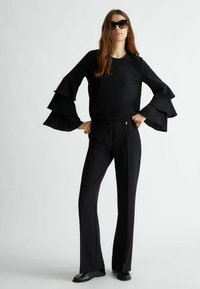 Top nero a maniche lunghe con ruches abbinato a pantaloni neri su misura. Il modello indossa occhiali da sole scuri. Texture liscia e design fluido.