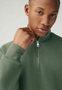 Sweat à zip vert en tissu texturé avec un motif en losange. Présente un col montant et des poignets côtelés. Détail de fermeture éclair argentée.