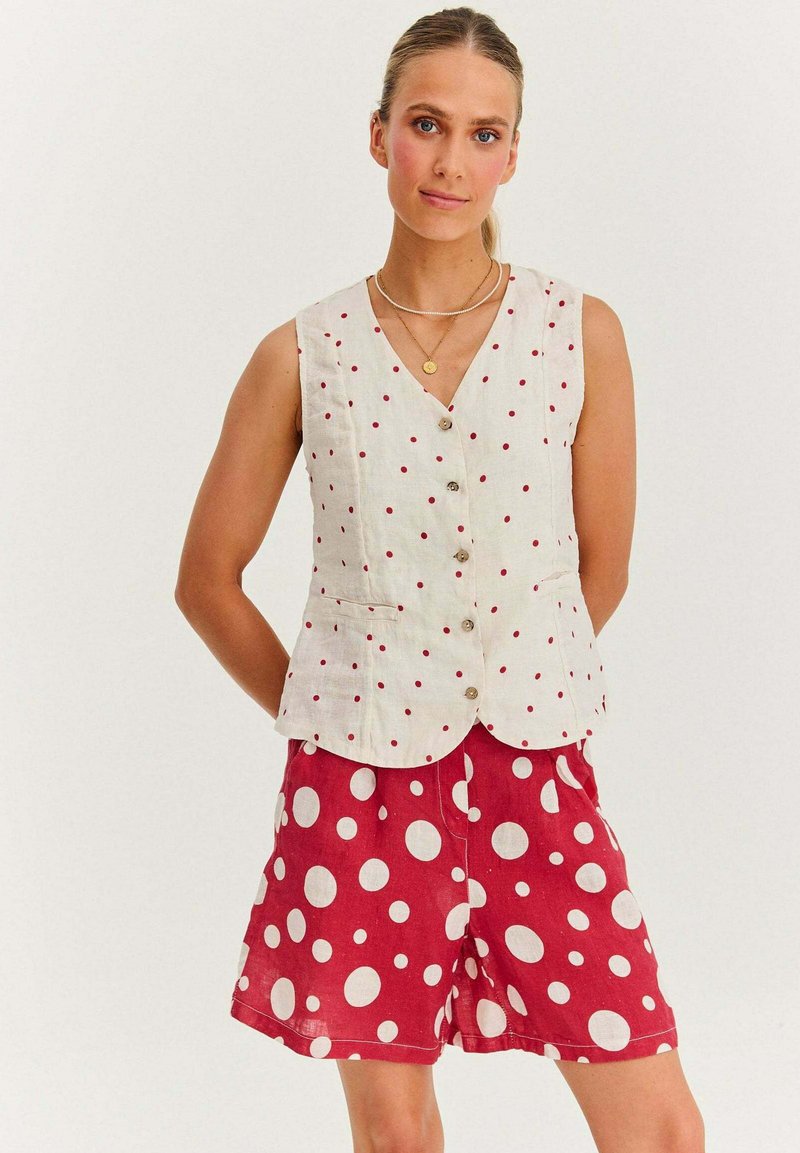 Biała lniana kamizelka w czerwone kropki, dekolt w serek i z przodu z zapięciem na guziki, w zestawie z czerwonymi szortami w duże białe kropki.