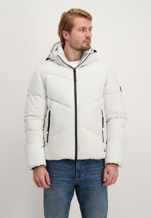 Witte pufferjack met een capuchon, ritssluiting, zwarte accenten en een geometrisch gestikt patroon aan de voorkant. Draagt met blauwe jeans.