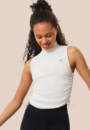 Weißes, geripptes Tanktop mit hohem Kragen, seitlichem Raffung mit Kordelzügen und kleinem Logo auf der Brust. Kombiniert mit schwarzen Leggings.