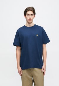 Temno modra bombažna majica s kratkimi rokavi in ribastim okroglim izrezom. Ima majhen zlat logotip na prsih.