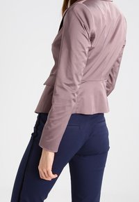 Femme portant un blazer ajusté mauve et un pantalon bleu marine avec une bande latérale noire, vue de dos sur un fond blanc.