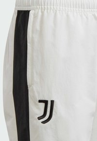 Witte sportieve broek gemaakt van soepel materiaal, met zwarte zijstrepen en een opvallend zwart geborduurd logo op de voorkant.