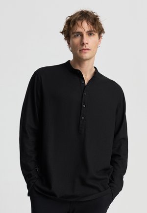 Giovane uomo con capelli mossi che indossa una maglia henley nera a maniche lunghe con bottoni, in piedi davanti a uno sfondo chiaro e uniforme.