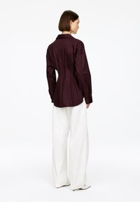 Woman facing away wearing a fitted dark purple long-sleeve shirt and wide white pants with white pointed heels.