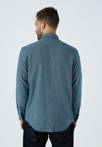 Mand med kort hår og skæg, iført en blå mønstret langærmet skjorte og mørke jeans, vendt væk mod en ensfarvet baggrund.