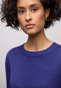Pull à col rond bleu profond, texture tricotée, coupe ajustée avec des détails côtelés au niveau de l'encolure. Tissu lisse, silhouette près du corps.