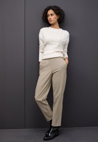 Pull en tricot torsadé crème associé à un pantalon beige à jambes tapered. Le mannequin porte des bottines noires. L'arrière-plan est gris foncé.