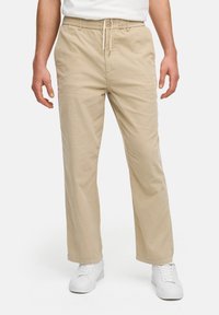 Pantalones de algodón beiges con cintura elástica y cordón, con un corte relajado y bolsillos laterales, combinados con zapatillas blancas.