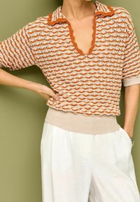 Korte mouwen trui van breiwerk met een beige en oranje zigzagpatroon, gegolfde halslijn en ribbelige beige zoom. Gemaakt in combinatie met losse witte broek.