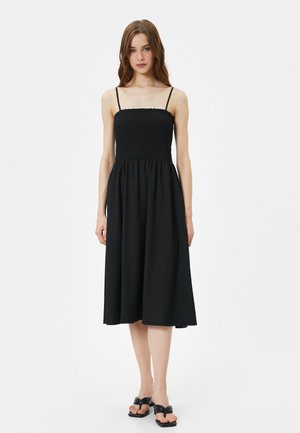 Robe midi noire avec un corsage smocké, des fines bretelles et une jupe fluide. Portée avec des sandales noires à enfiler, sur fond blanc.