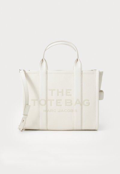 Sac fourre-tout en cuir blanc avec le texte "LE SAC FOURRE-TOUT" imprimé en clair. Design structuré, poignées supérieures doubles, bandoulière amovible.