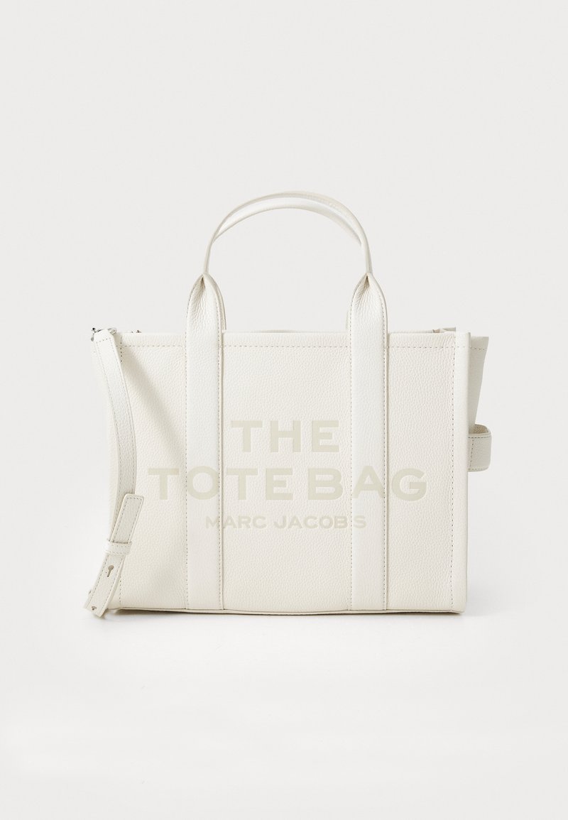 Valkoinen nahkainen kassi, jossa on "THE TOTE BAG" -teksti vaalealla painatuksella. Rakenteellinen muotoilu, kaksinkertaiset yläkahvat, irrotettava olkahihna.