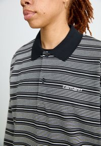 Polo a righe bianche e nere con colletto nero, due bottoni e logo ricamato "Carhartt" sul lato sinistro del petto.