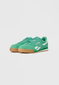 Zaļas zamšādas kedas ar baltām akcentiem, apaļa purngala forma, plakans gumijas zole un saitēm veidots dizains. Malā ir zils Reebok logotips.