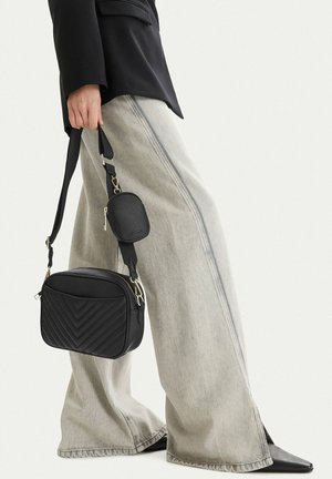 Persona vistiendo jeans de pierna ancha gris claro, zapatos negros puntiagudos y blazer negro, sosteniendo un bolso bandolera negro acolchado con un pequeño monedero.