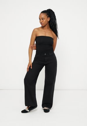 Femme aux longs cheveux tressés portant un haut noir sans bretelles, un pantalon noir à jambes larges et des ballerines noires, debout devant un fond blanc.