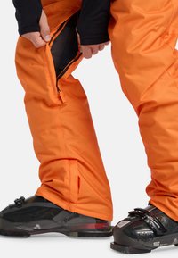 Ruky upravujú zips vetrania na jasne oranžových lyžiarskych nohaviciach, ktoré sú oblečené cez čierne lyžiarske čižmy s prackami.