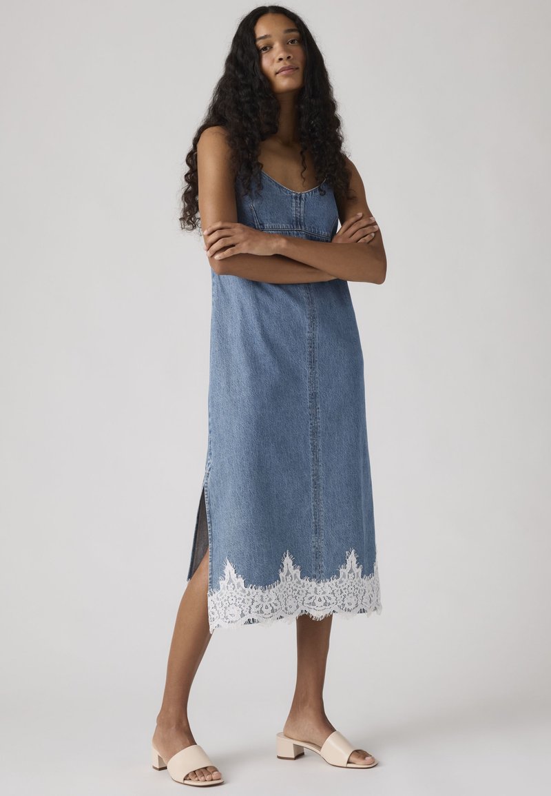 Frau mit langen lockigen Haaren steht mit verschränkten Armen, trägt ein ärmelloses blaues Denim-Midikleid mit weißer Spitzensaum und beige Sandalen mit klobigem Absatz.