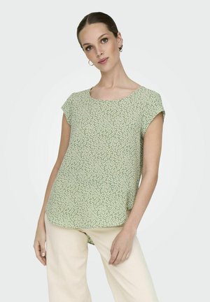 Green short-sleeve blouse with a rounded neckline, featuring a textured floral pattern and a relaxed fit, complemented by ivory pants.