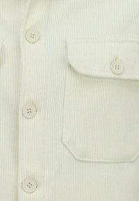 Roomwit geribd shirt met twee grote borstzakken, vier knopen aan de voorkant en een zachte textuur.