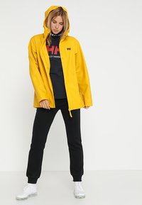 Geel waterdichte jas met capuchon, voorzien van een rits en drukknoopsluiting, gelaagd over een zwarte sweatshirt en zwarte broeken. Witte sneakers.