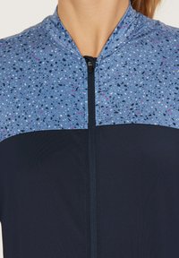 Veste de sport bleue avec un motif tacheté sur la partie supérieure, une section inférieure en bleu marine uni et une fermeture éclair à l'avant. Tissu texturé.